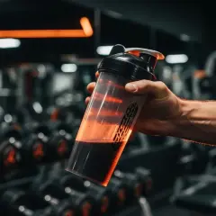 Main d'un homme tenant un shaker rempli de liquide sombre, dans une salle de sport avec des haltères en arrière-plan. La whey sert à la récupération musculaire.