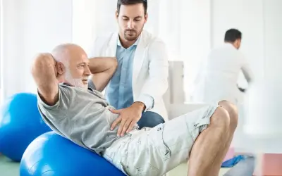 Un kiné guide un homme âgé dans sa rééducation périnéale sur un ballon de gym.