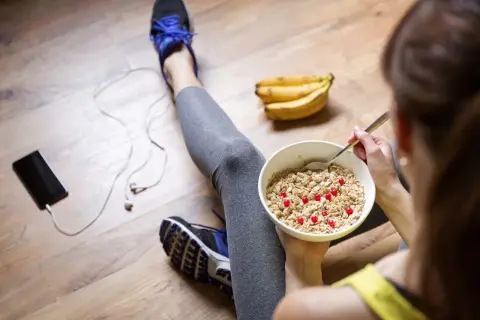 Après le sport, une femme mange un bol de flocons d'avoine avec des baies rouges. Elle est assise sur le sol en bois, à côté de ses chaussures de sport, d'un téléphone et d'écouteurs. Elle suit un régime pour maigrir.