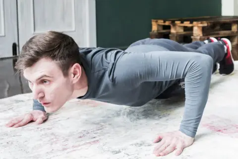 Un homme en tenue de sport fait des pompes tous les jours sur un tapis. Il est concentré et déterminé.