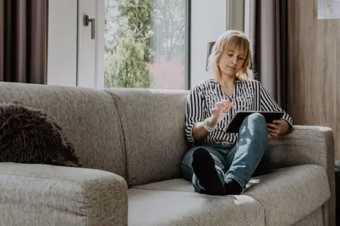 Femme blonde sur un canapé gris, consultant une tablette. Elle semble chercher un avis sage-femme, peut-être pour une grossesse.