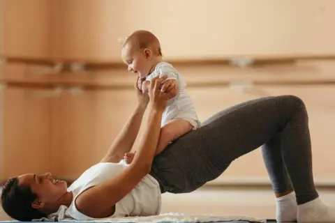 Une mère fait des exercices pour renforcer son périnée après accouchement, soulevant son bébé joyeux.