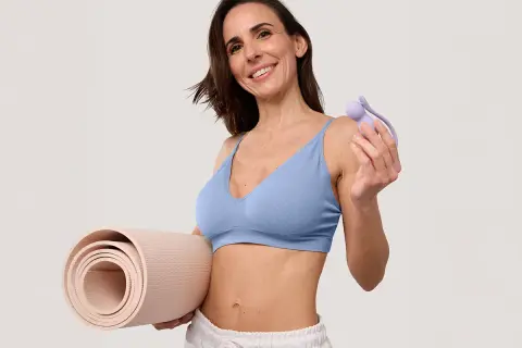 Une femme souriante tient un accessoire pour muscler son périnée à 60 ans et un tapis de yoga.