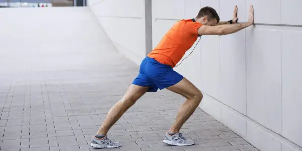 Un homme en tenue de sport fait un étirement mollet contre un mur blanc.