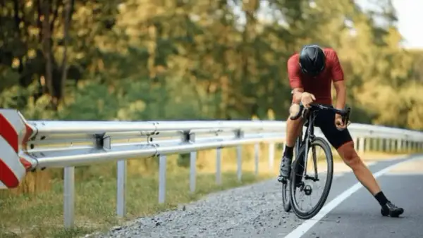 Cycliste en tenue de sport, casque noir, s'appuyant sur son vélo sur le bord de la route. Il semble fatigué, peut-être après avoir fait du sport à jeun.