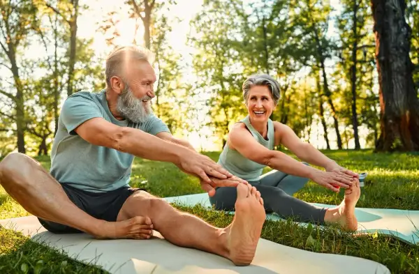 Couple souriant pratiquant le qi gong en plein air, étirant leurs jambes sur des tapis de yoga dans un parc ensoleillé.